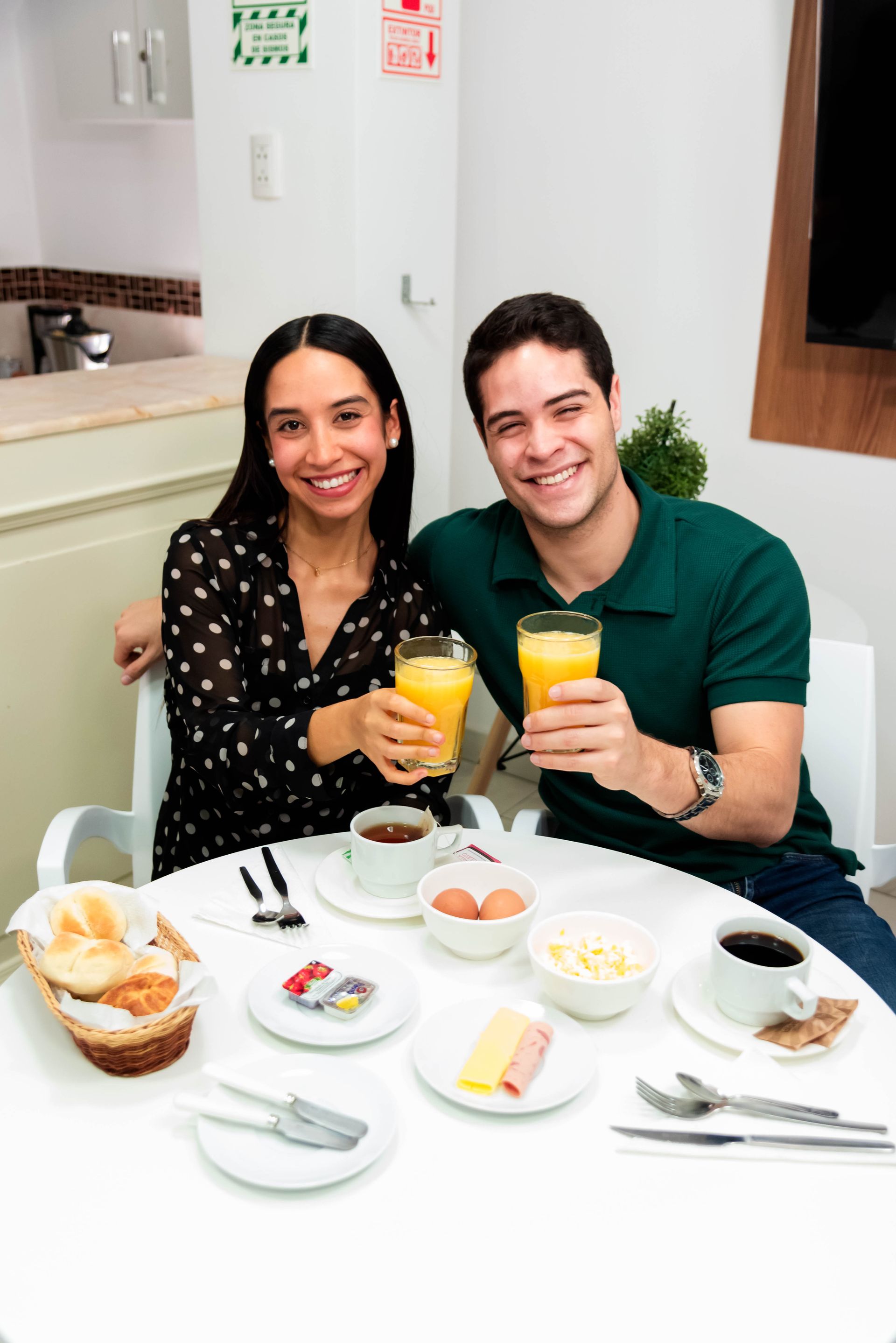 Una pareja brinda con jugo de naranja en la mesa del desayuno. Sonríen. La mesa tiene comida y café, y está en el interior.