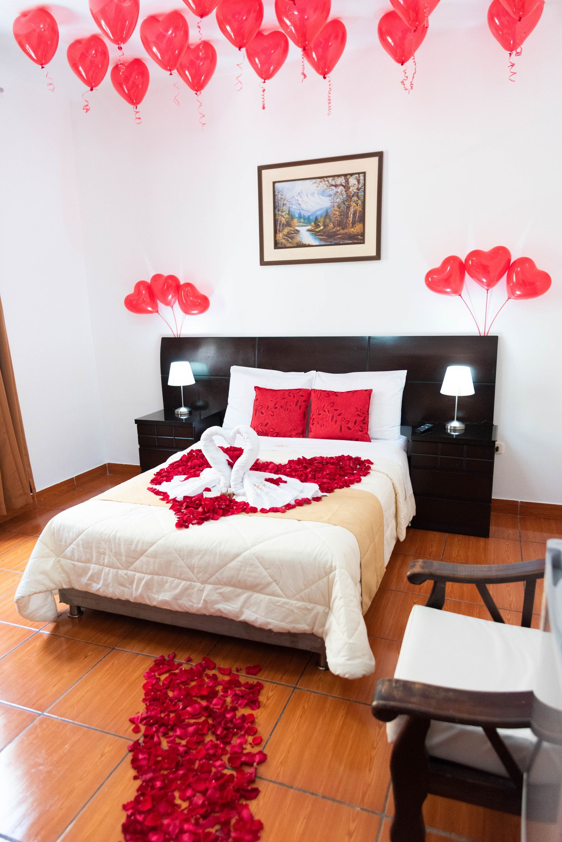Habitación de hotel romántica decorada con globos de corazón rojo y pétalos de rosa en la cama y el suelo.