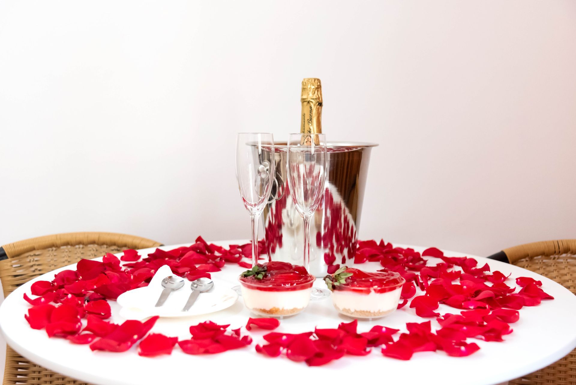 Mesa romántica con champán, postres, pétalos de rosa y cubiertos.