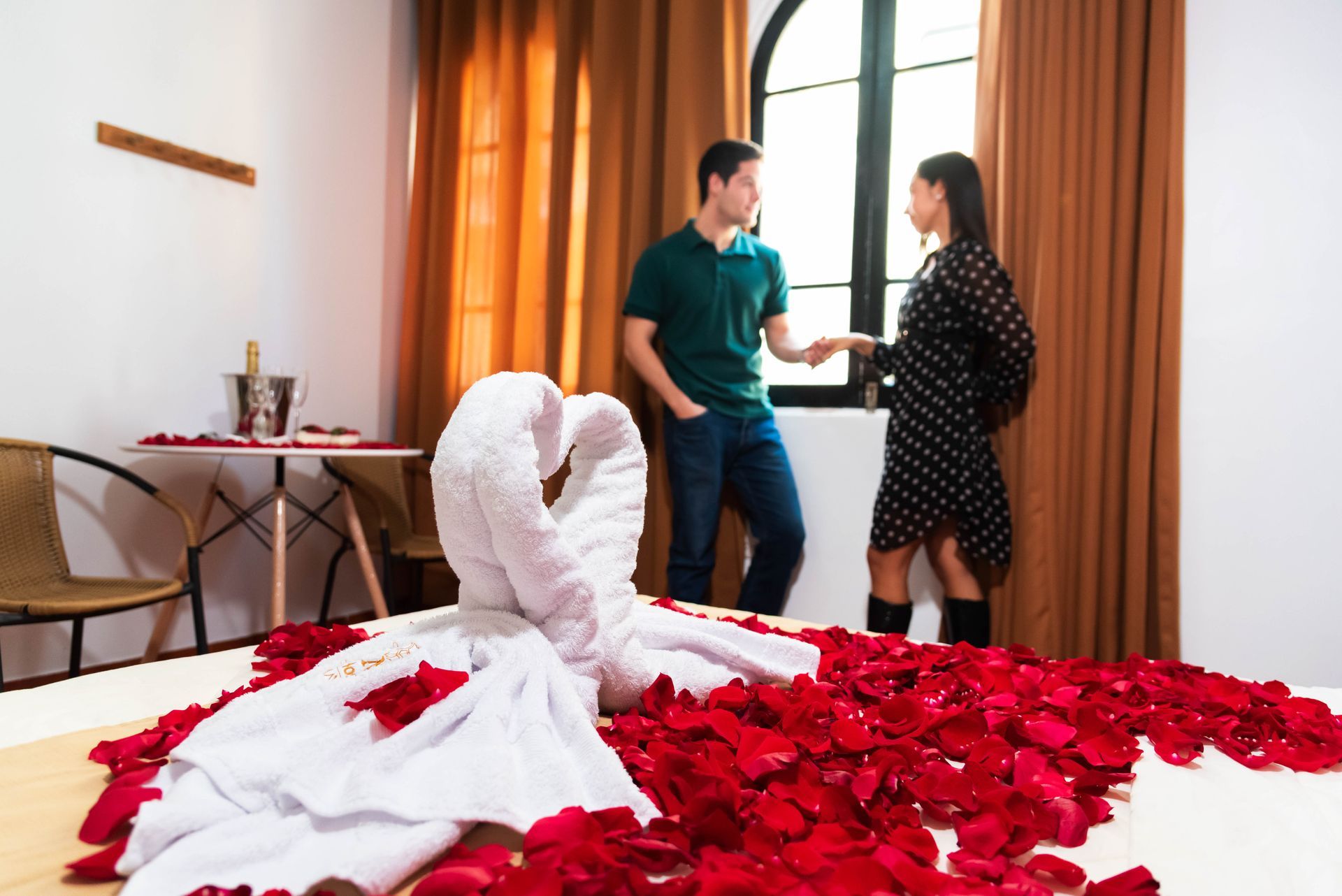 Habitación de hotel romántica con pareja. Pétalos de rosa rojos sobre la cama con una toalla. Pareja de pie, sonriendo.