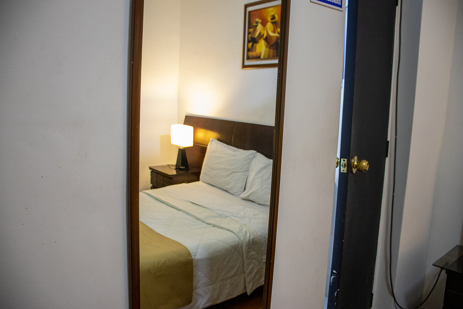 Un espejo refleja una pequeña habitación de hotel con una cama, una mesita de noche y arte enmarcado.