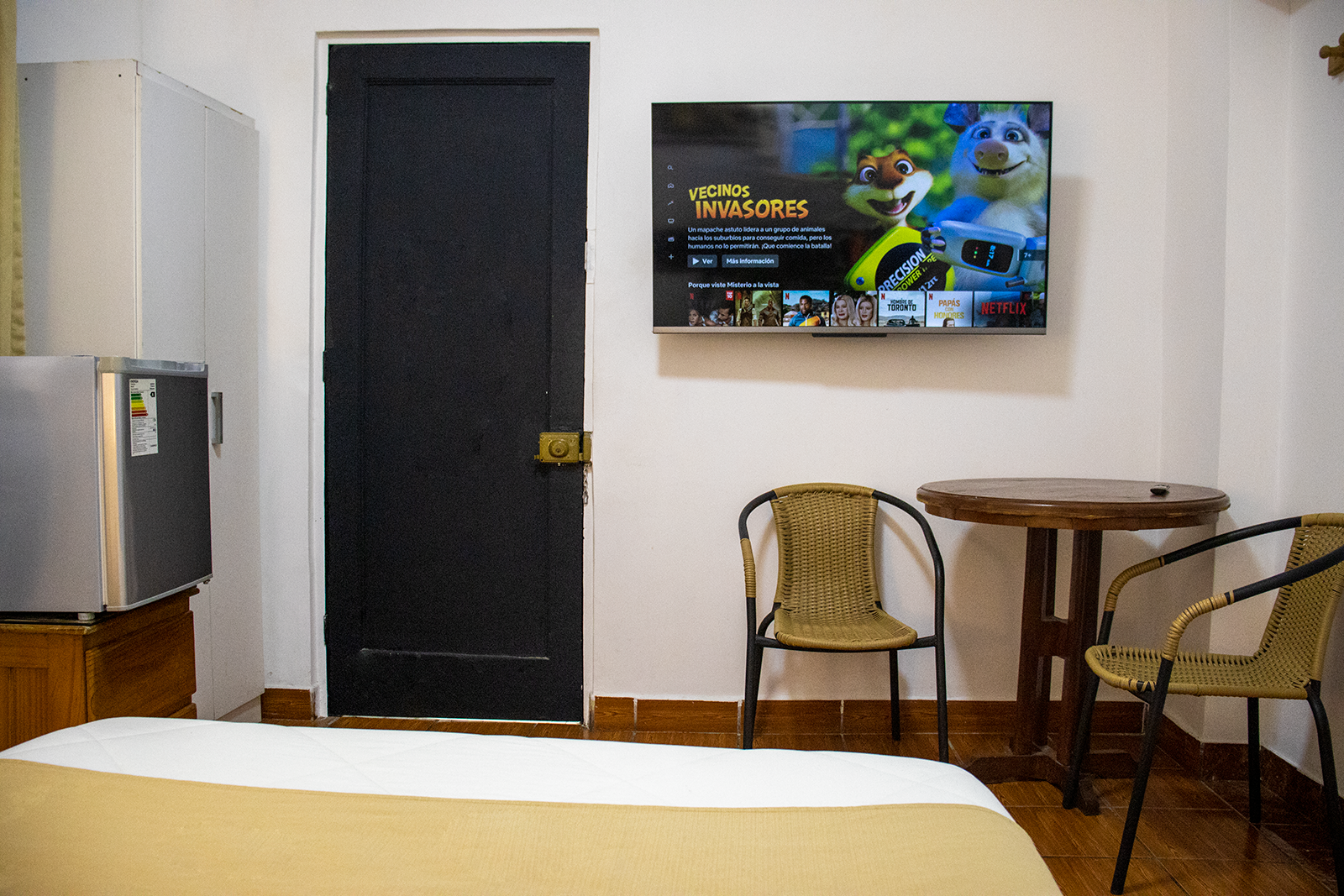 Habitación pequeña con cama, televisor, sillas y mesa. También incluye nevera y puerta negra.