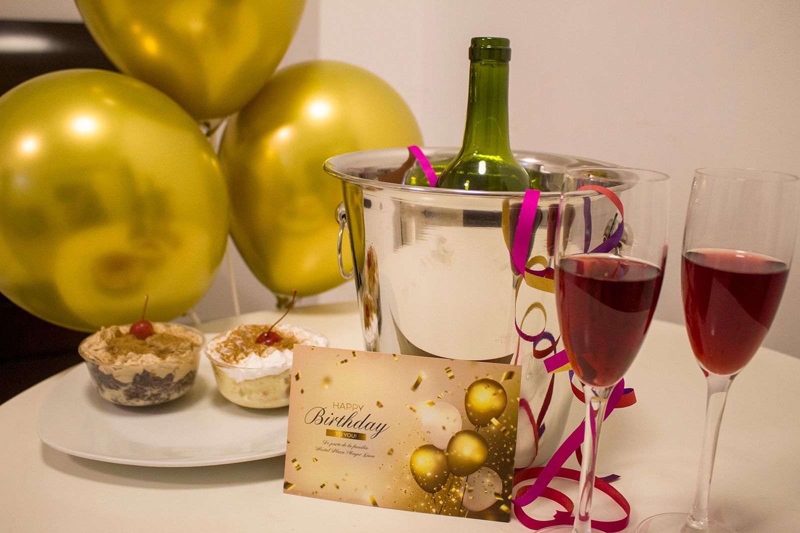 Celebración de cumpleaños con globos, cupcakes, champán y una tarjeta en una mesa.