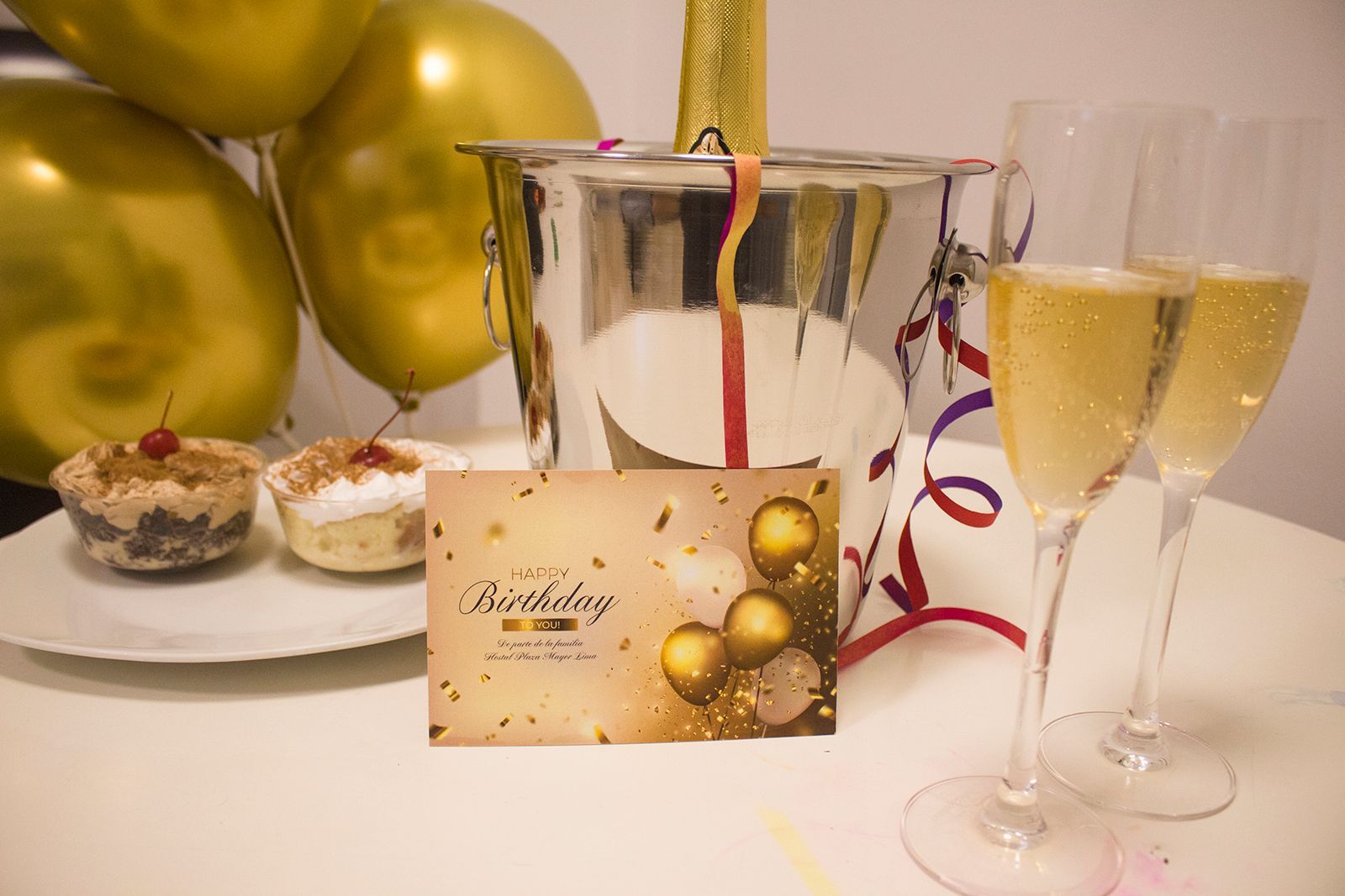 Celebración de cumpleaños con champán, cupcakes, tarjetas, globos y un balde.