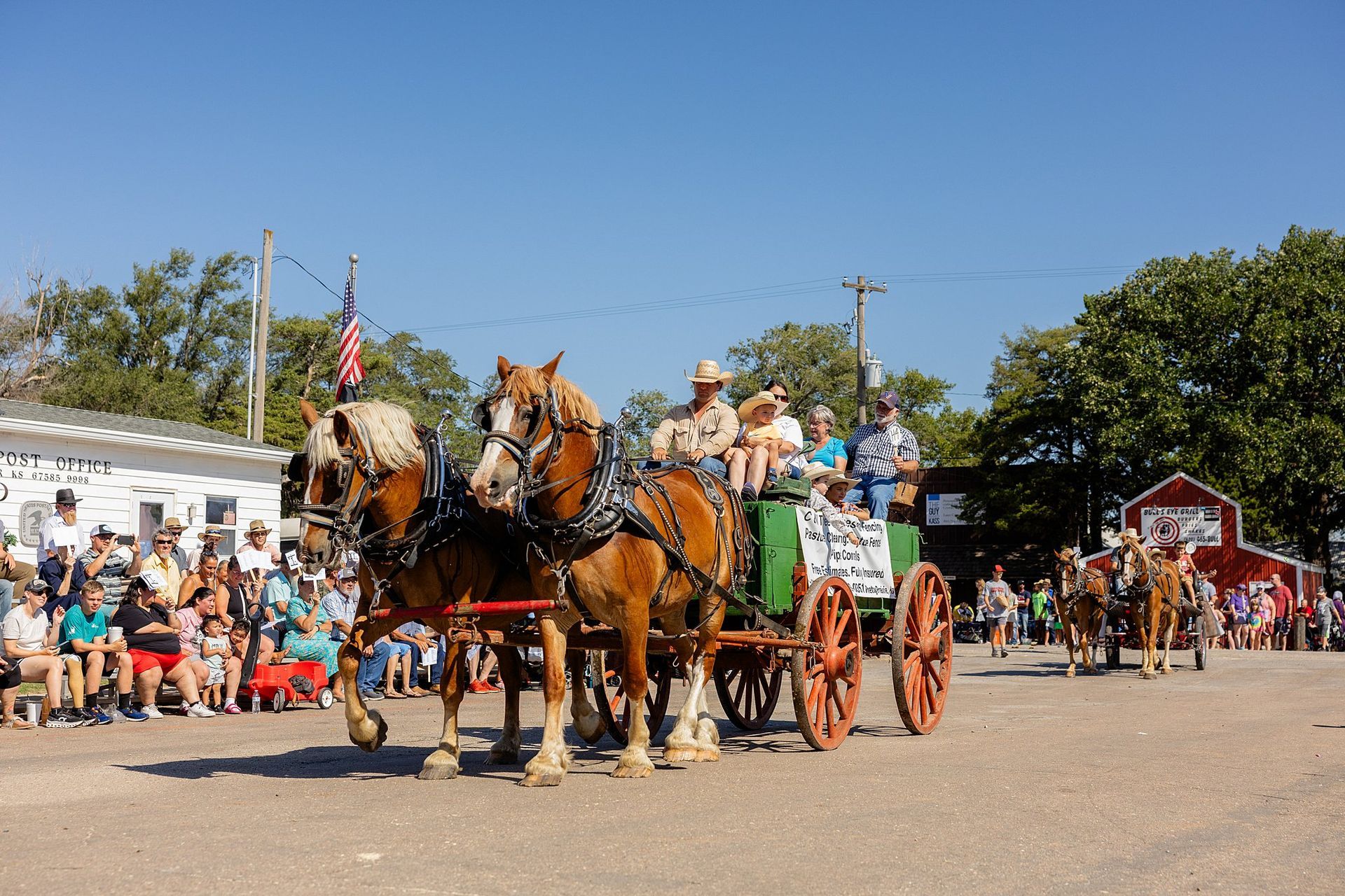 Two draft horses pull a green wagon with people in a parade, spectators line the street on a sunny day.