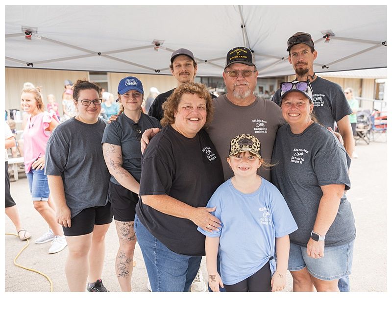 Group of people posing outdoors under a tent, smiling. People in matching t-shirts, some wearing hats.