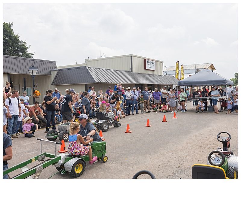 Children on toy tractors competing in a race with spectators and a tent in the background.