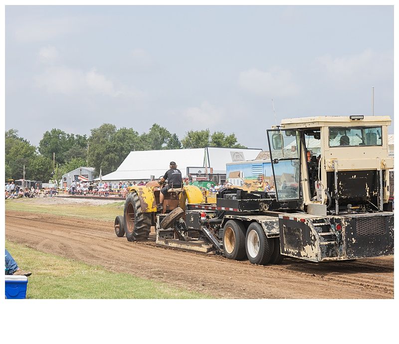 Yellow tractor pulling a sled on a dirt track, with a large beige truck and spectators in the background.