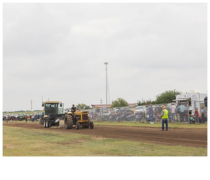 Tractor pulling competition on dirt track; spectators, cloudy sky.