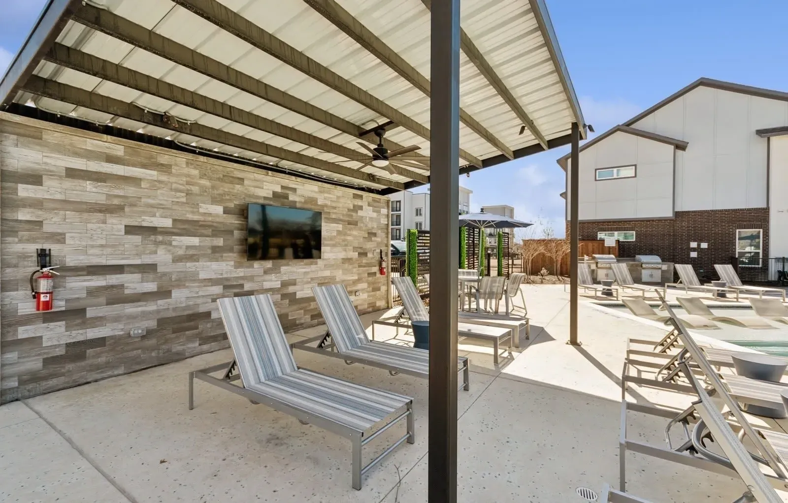 Outdoor pool deck with striped lounge chairs under a metal canopy and a wall-mounted TV. See what makes our pet-friendly apartments in Rowlett, TX a welcoming place to call home—schedule a tour.