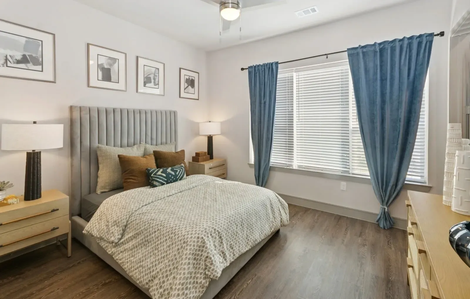 Bedroom in apartment with upholstered headboard, two nightstands, dresser, and blue curtains by a window. Relax and recharge at luxury apartments in Rowlett, TX with pool and gym—schedule a tour to see it all.