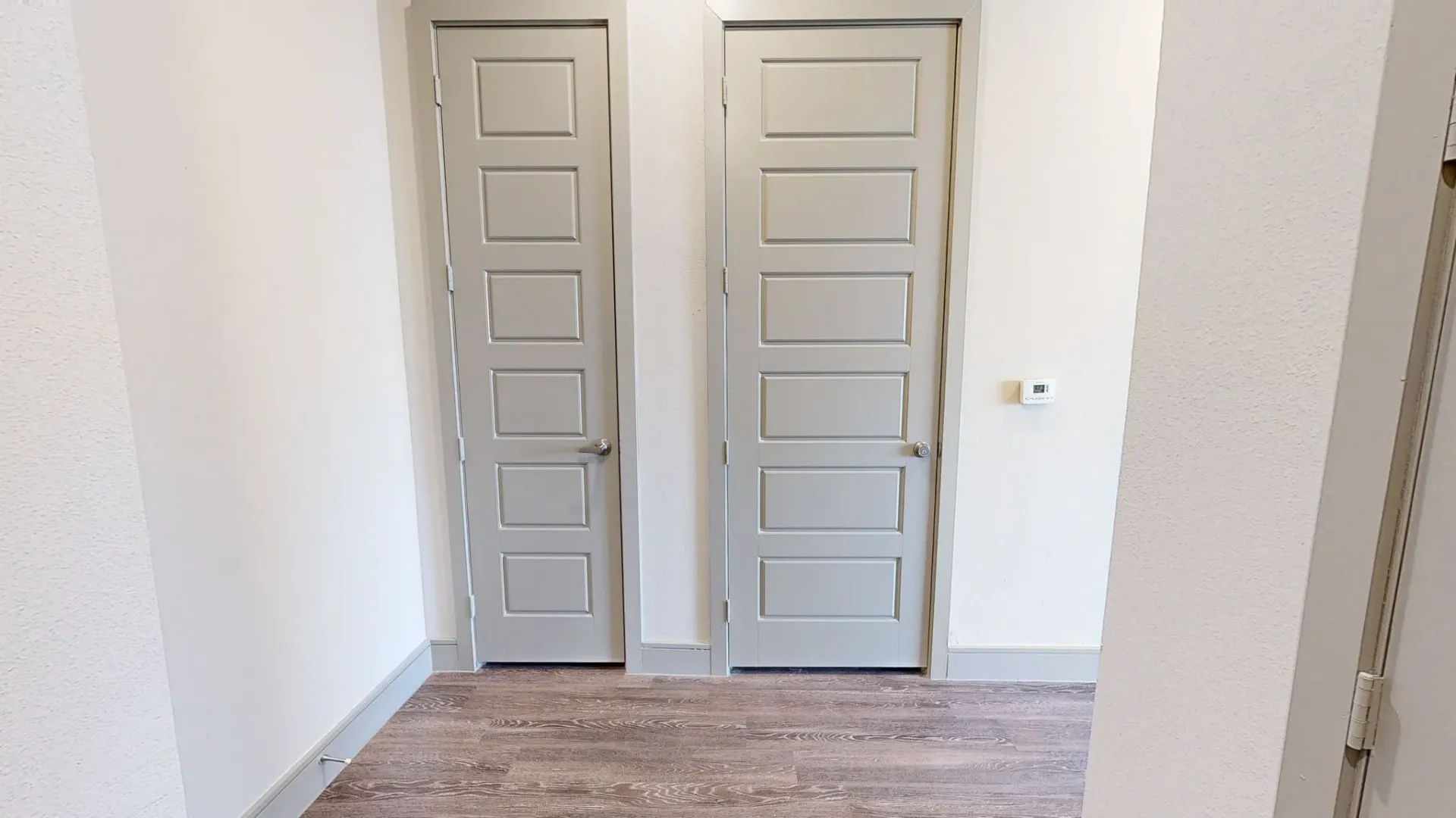 Interior hallway with two paneled gray doors, light walls, and wood-like flooring. Experience refined comfort at luxury apartment homes near Pecan Grove Park—book your tour today.