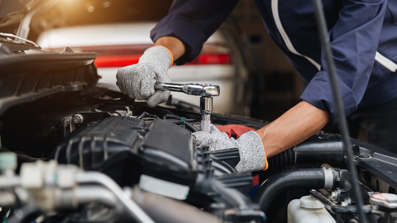 Mechanic in blue jumpsuit uses a wrench on a car engine, wearing gloves.