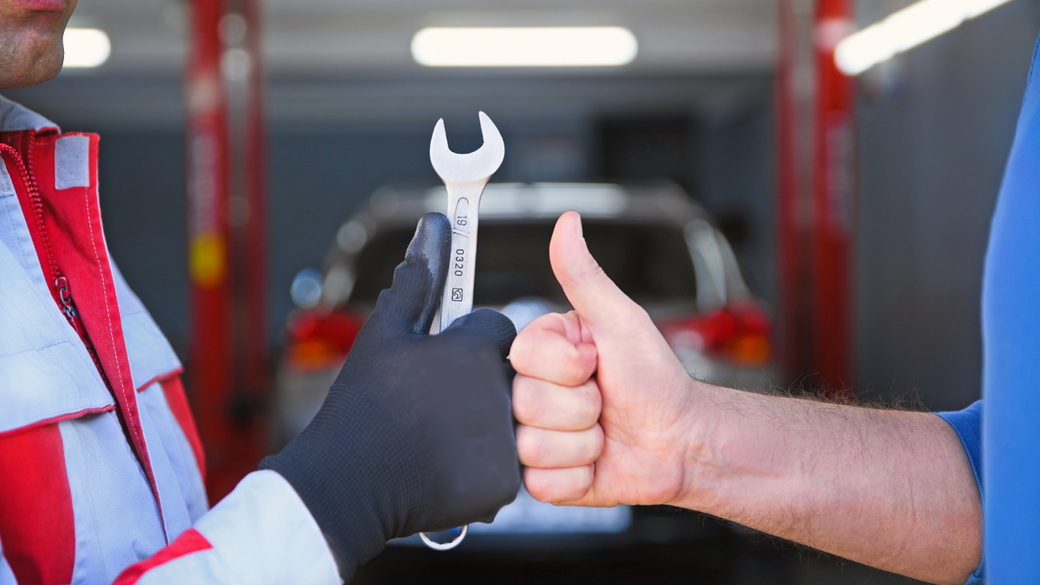 Mechanic in red and white suit hands a wrench to a person giving a thumbs-up in a garage.