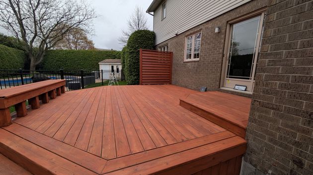 Terrasse en bois teinté en rouge, prolongeant une maison en briques et donnant sur un jardin clôturé avec piscine.