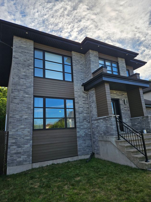 Maison moderne à deux étages avec des éléments en pierre grise et des touches de brun, de grandes fenêtres, des finitions noires et une petite cour avant avec des marches.