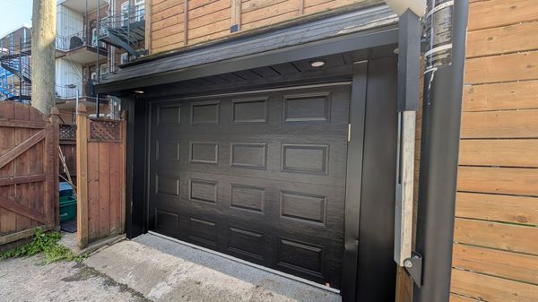 Porte de garage noire à panneaux texturés, à côté d'une clôture en bois et d'un bâtiment avec un escalier en métal.