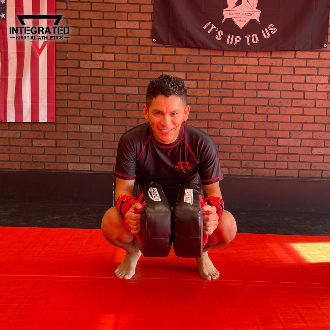 A man is squatting on a red mat holding a pair of boxing gloves.
