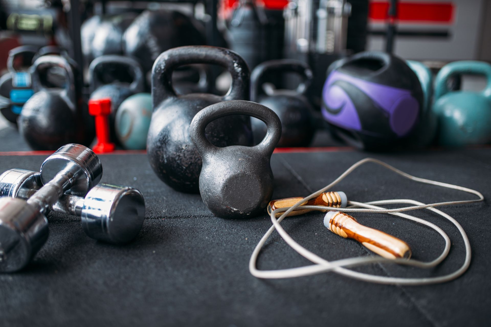 A jump rope , dumbbells , and kettlebells are on the floor in a gym.