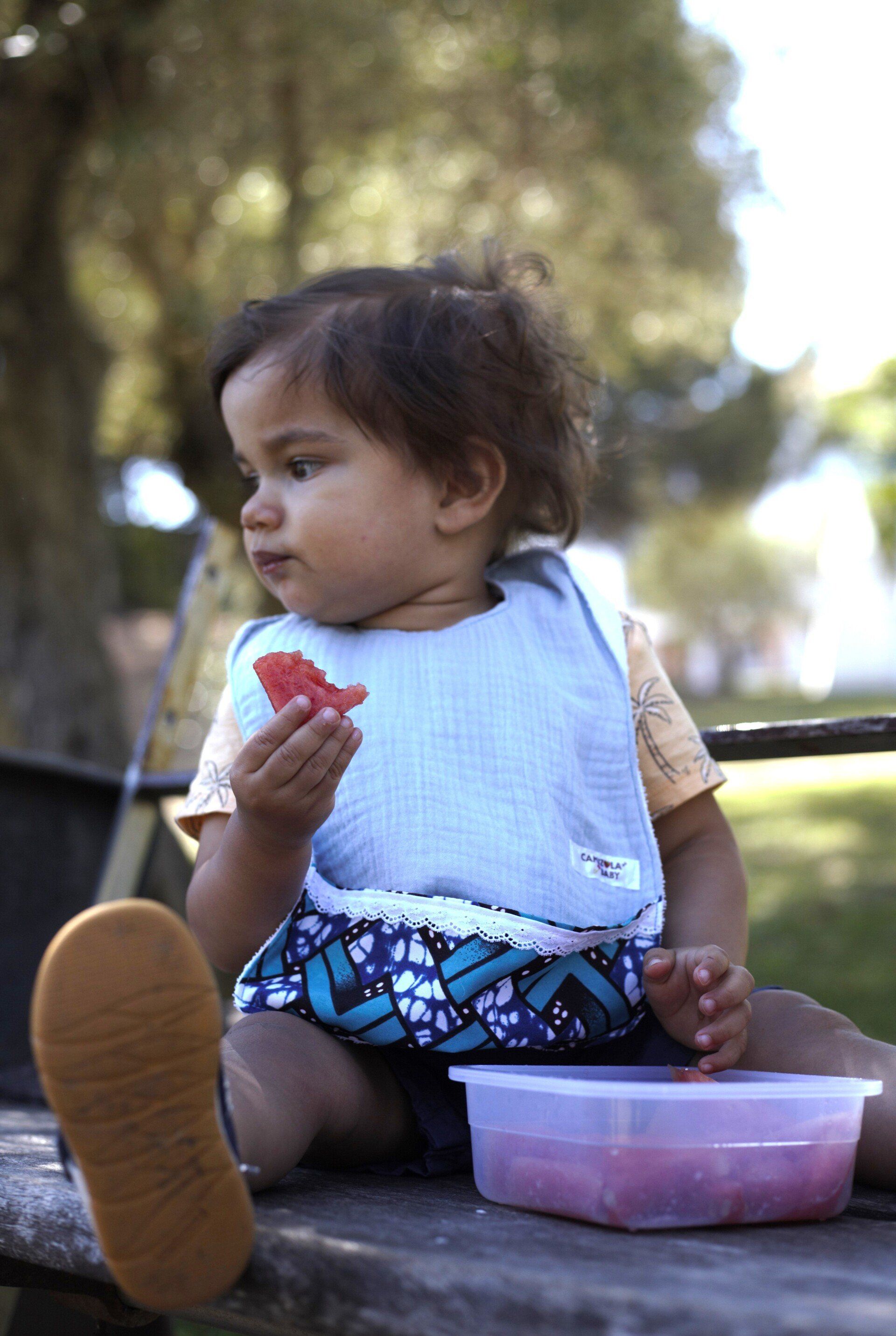 Um bebê está sentado em um banco comendo uma fatia de melancia.