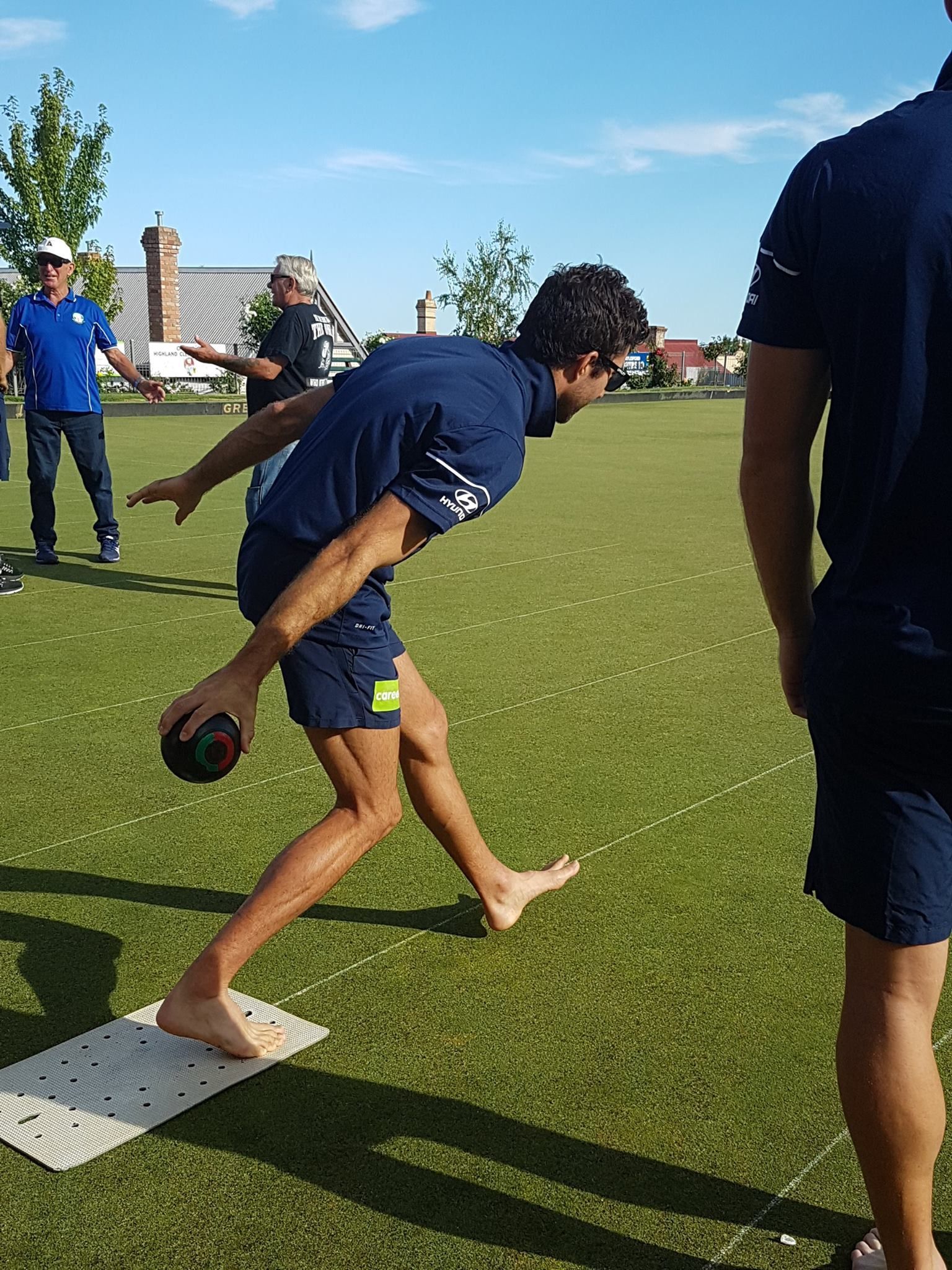 Young man on grass playing lawn bowls, wearing navy t-shirt and shorts.