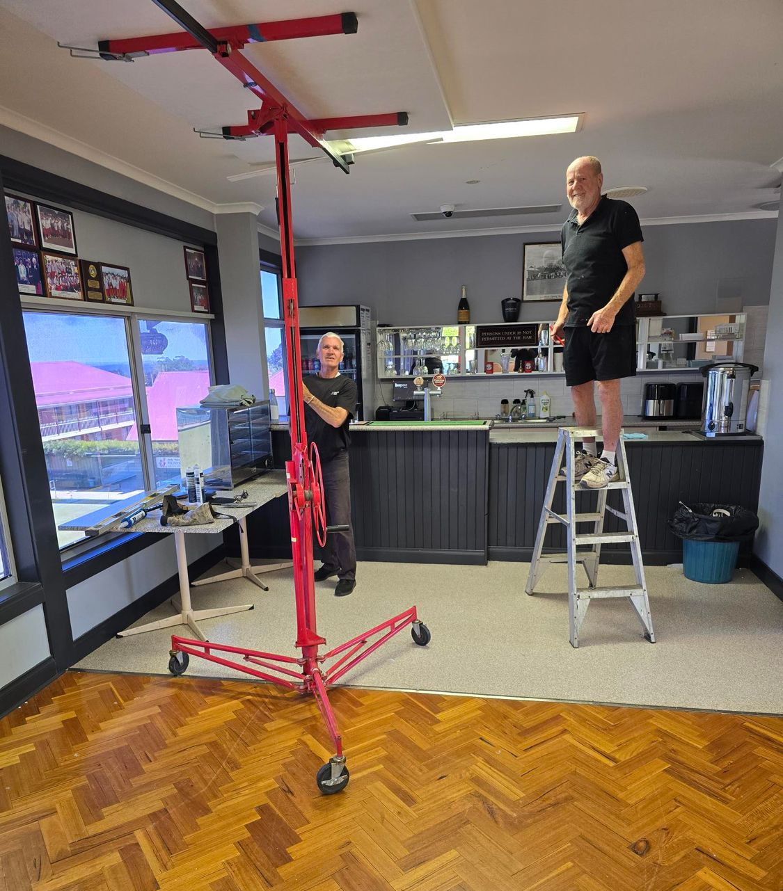 Man on ladder indoors wearing black polo shirt and shorts. Another man wearing black t-shirt and pants operating a plaster installing machine.