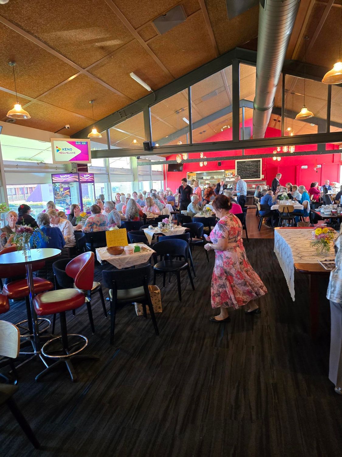 People in venue seated for meal. A lady with brown hair is in the front walking past in a floral dress and hair up.