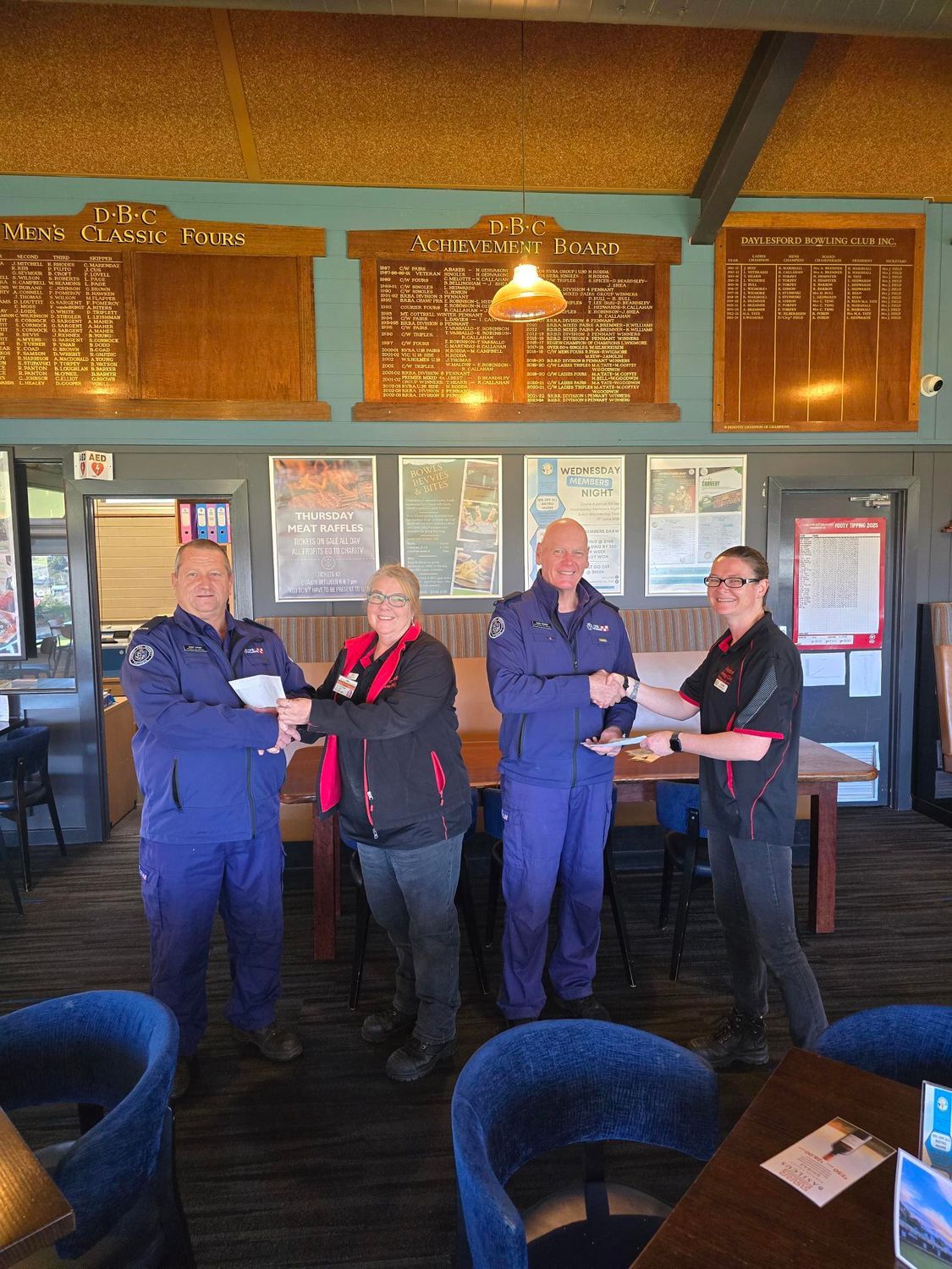 Two female staff members  dressed in black shaking hands with two male emergency services crew dressed in their blue uniform and presenting them with a cheque.