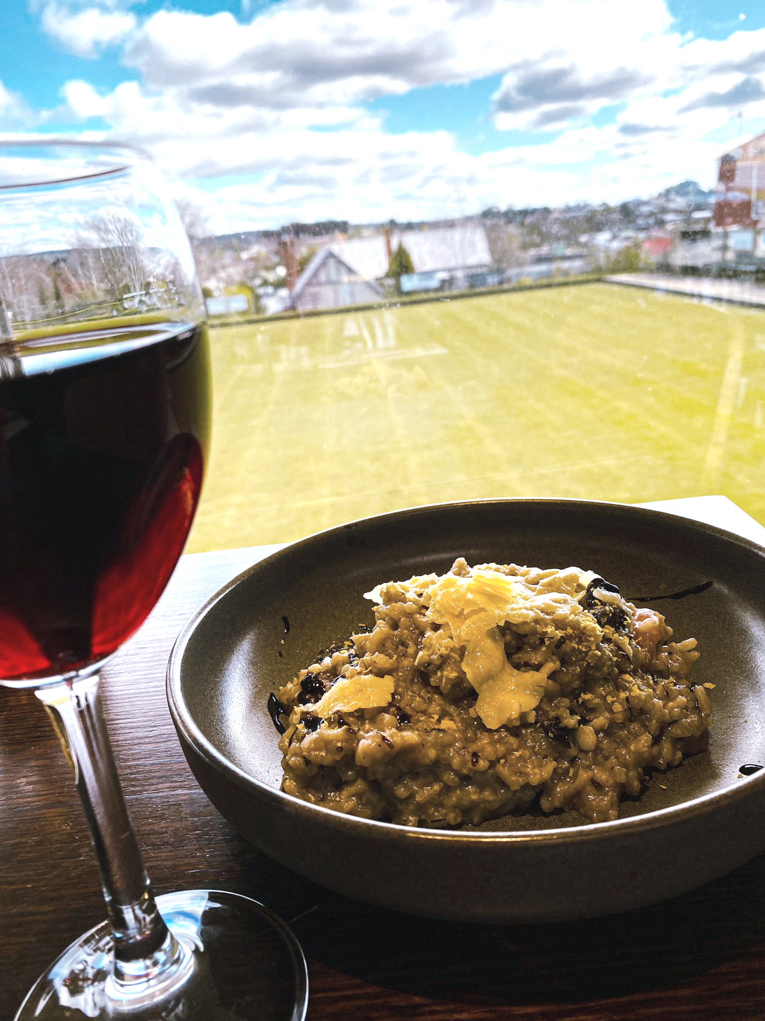 Brown bowl with risotto piled up with shavings of cheese. Glass of red wine to the left and window looking out to bowling green. There are houses in the background.
