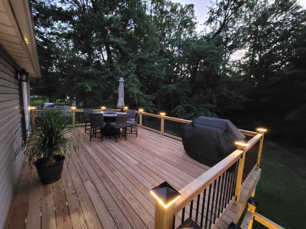 Wooden deck at dusk with dining table, grill, patio umbrella, and lit railings overlooking trees