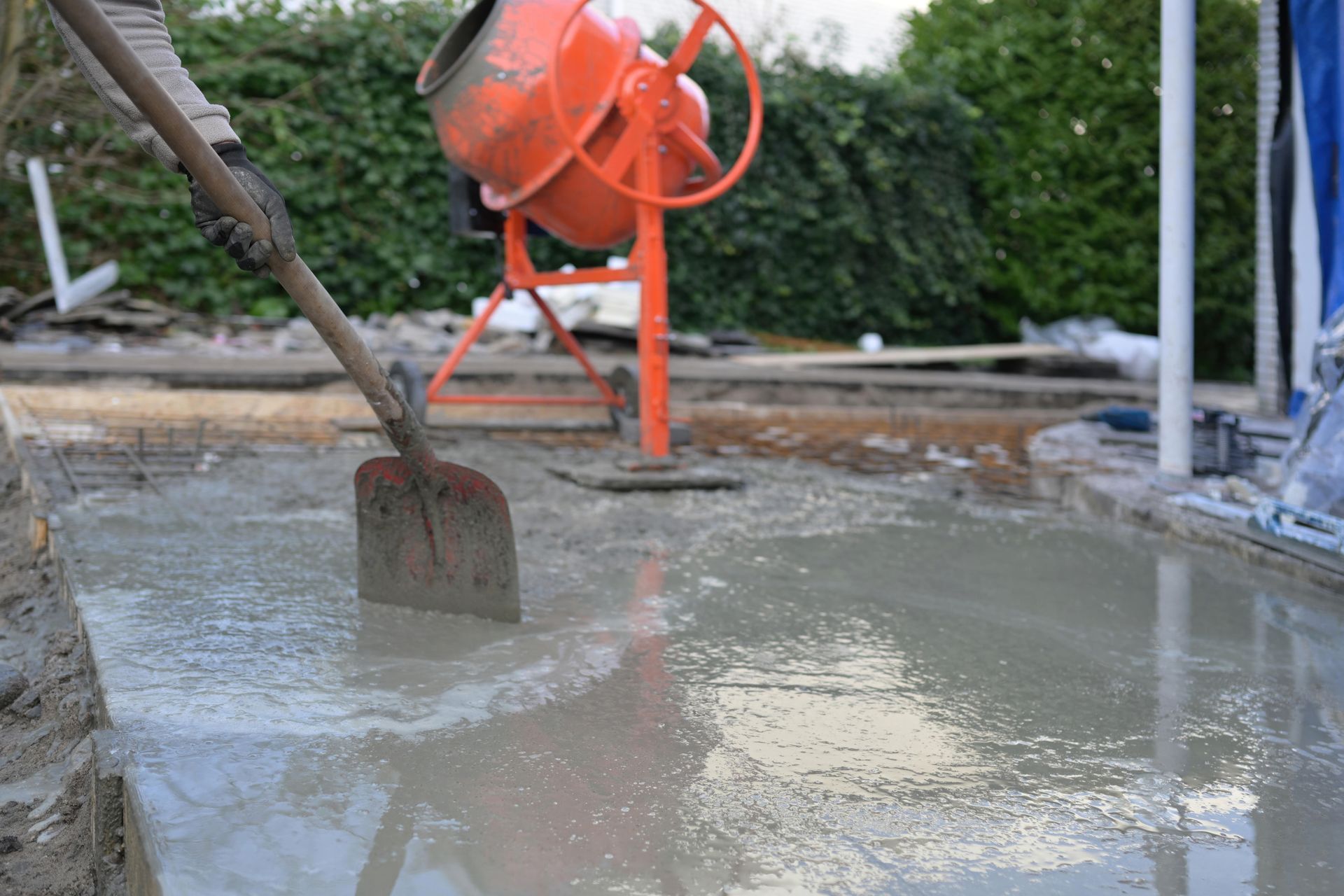 Worker smoothing wet concrete with a shovel, orange cement mixer in the background.