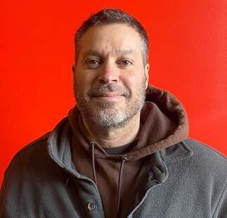 A man with a beard is wearing a gray jacket and a brown hoodie.