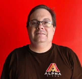 A man wearing glasses and a brown shirt is standing in front of a red background.
