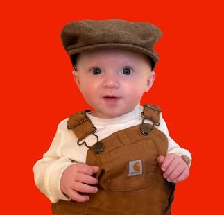 A baby wearing overalls and a hat on a red background