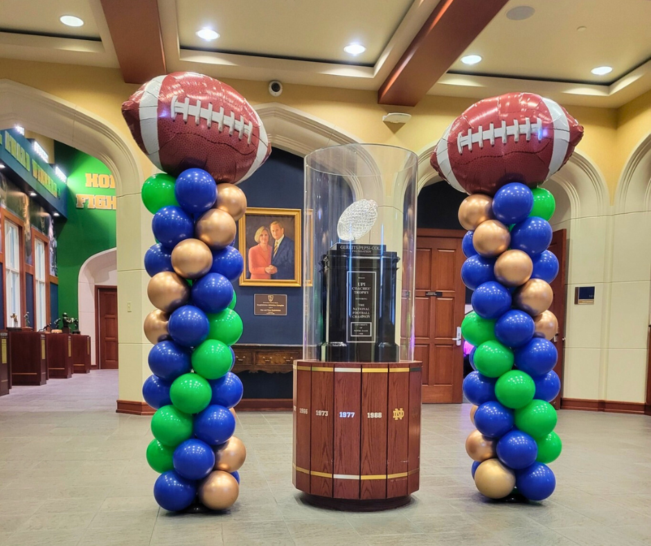 Two football shaped balloons are sitting next to each on balloon columns at ND next to the championship trophy