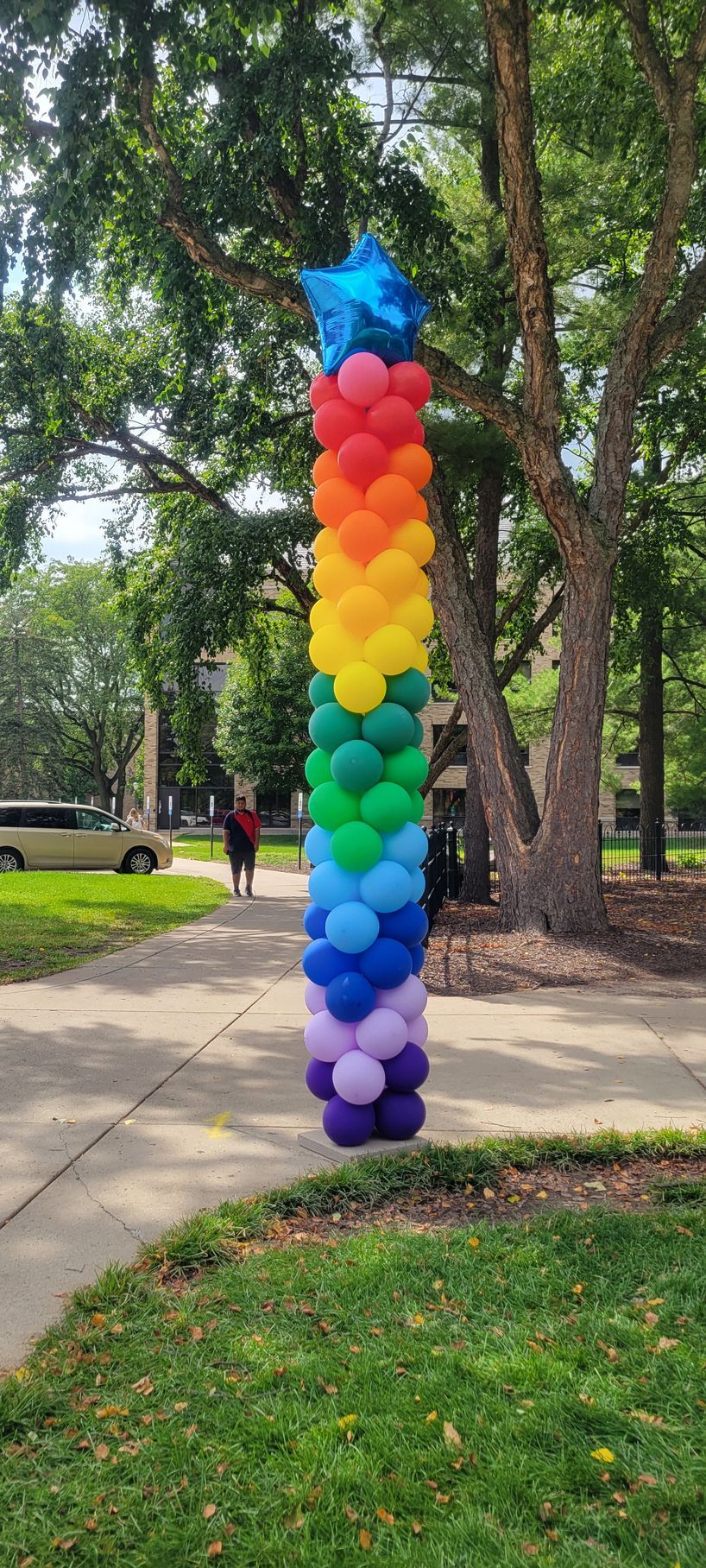 Rainbow balloon column with a blue star topper on a sidewalk, in front of a tree-filled background.