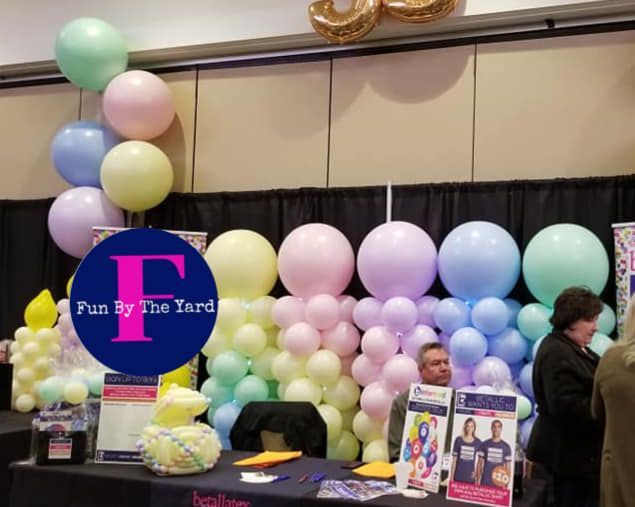A table with balloons and a sign that says fun by the yard. Tradeshow balloon columns.
