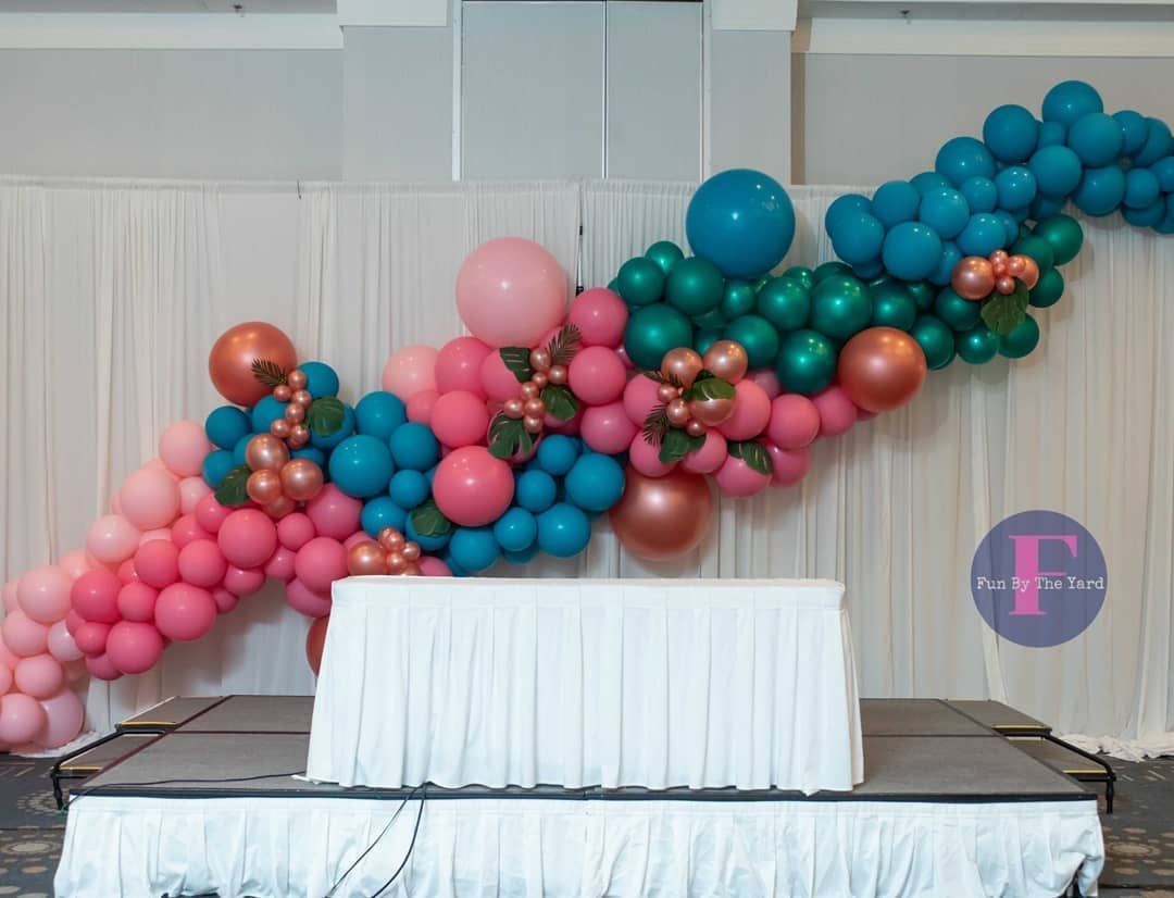 Stage decorated with a cascading balloon garland in pink, teal, and copper, above a white-skirted table.