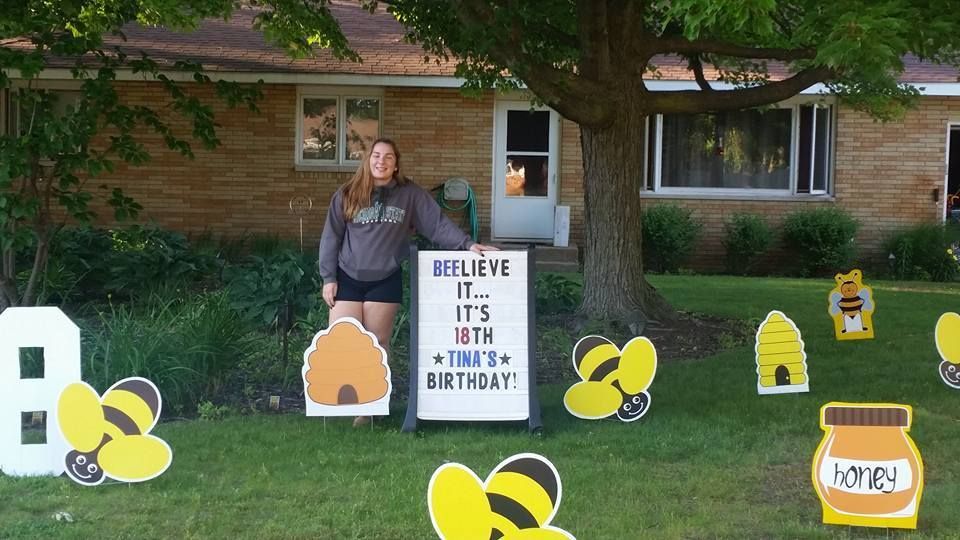 A woman is standing in front of a house with a sign that says believe it. Bee themed yard card.