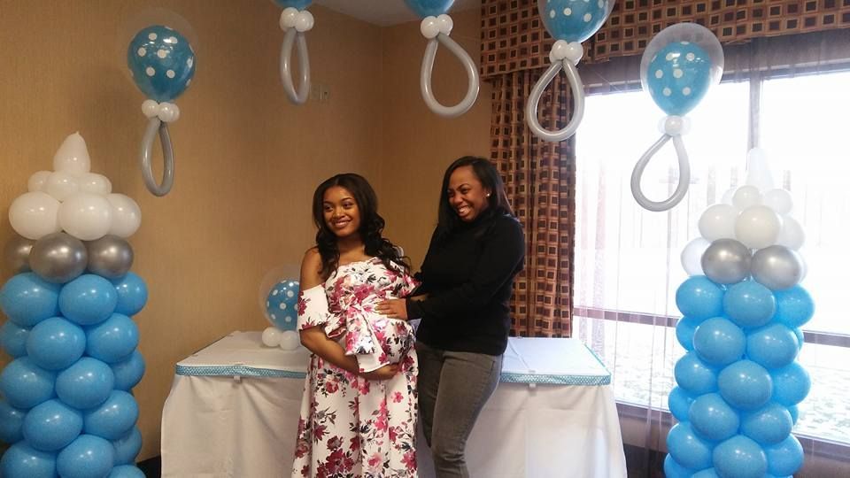 Two women are standing next to each other in a room decorated with balloons. Baby shower balloon bottles and pacifier arch