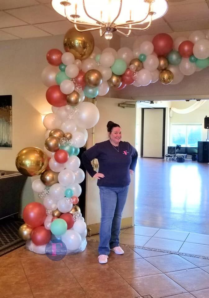 Woman poses near a balloon arch with gold, white, and coral balloons at an event entrance.