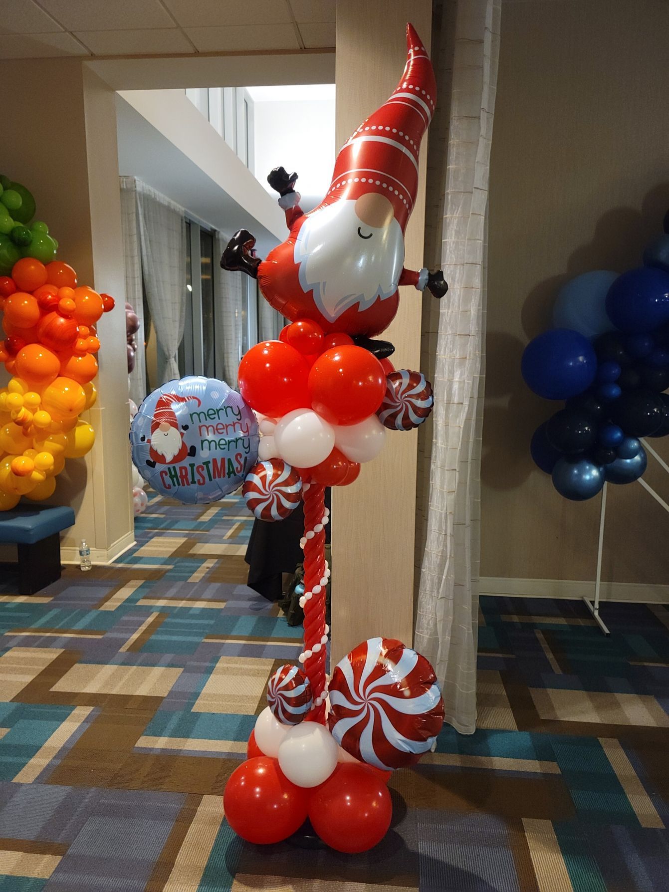 A balloon gnome is sitting on top of a candy cane in a hallway.