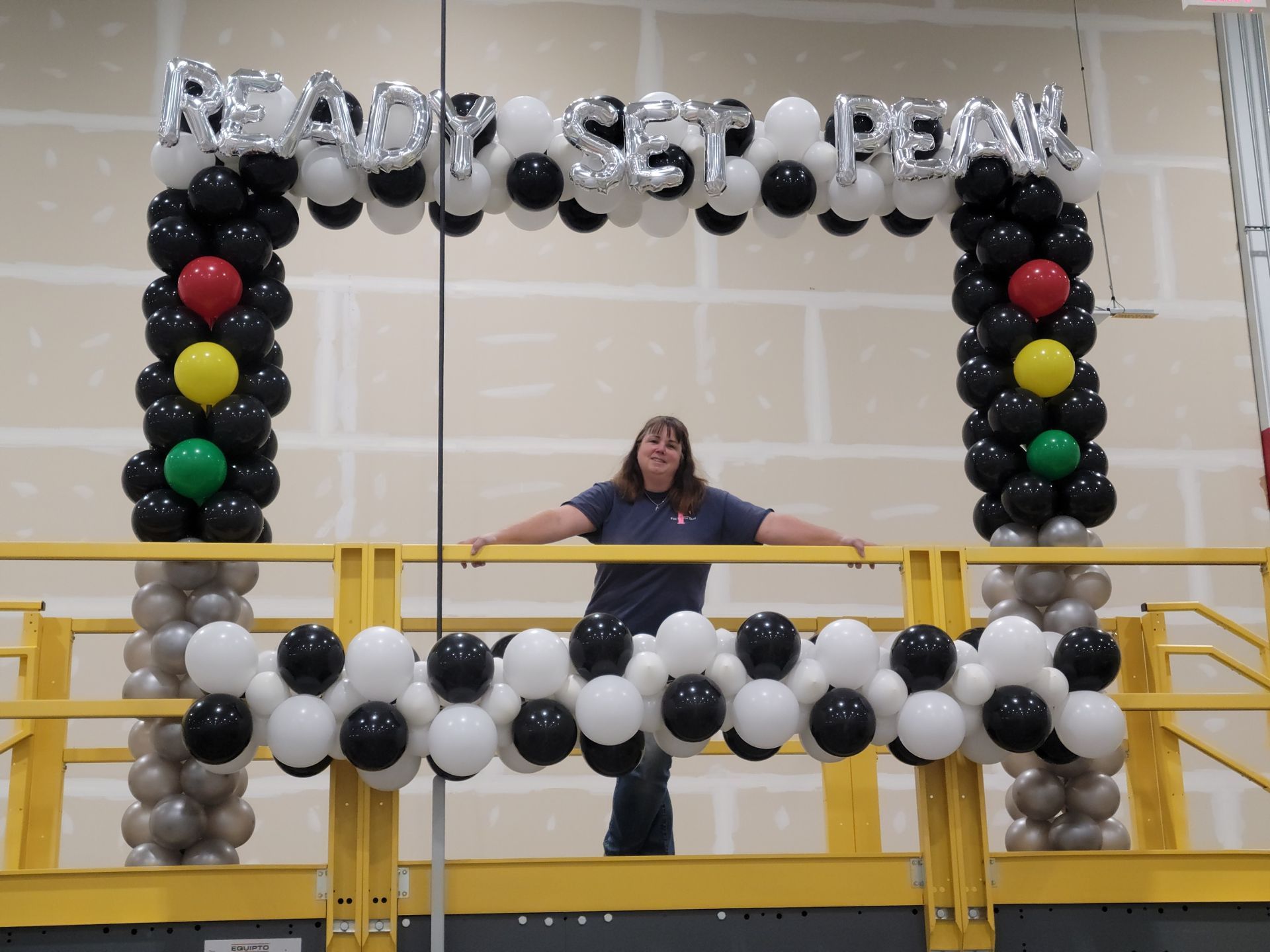 A woman stands in front of a balloon arch that says ready set peak at Amazon. Racecar theme.