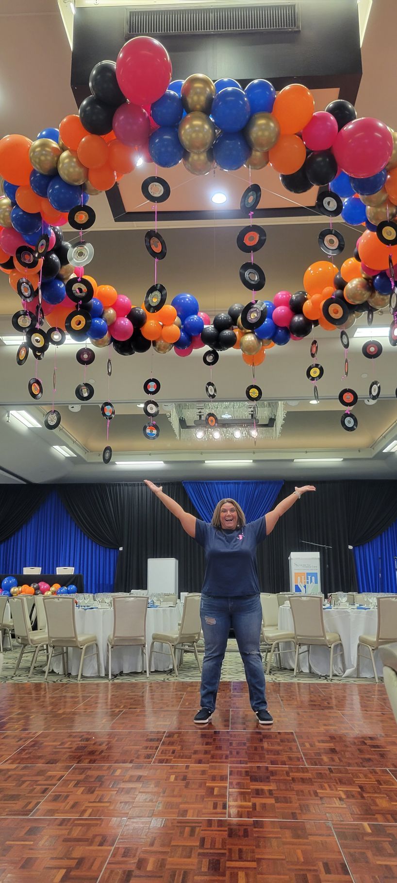 Woman with arms raised stands on a dance floor. Balloons and vinyl records decorate the ceiling for a celebration.