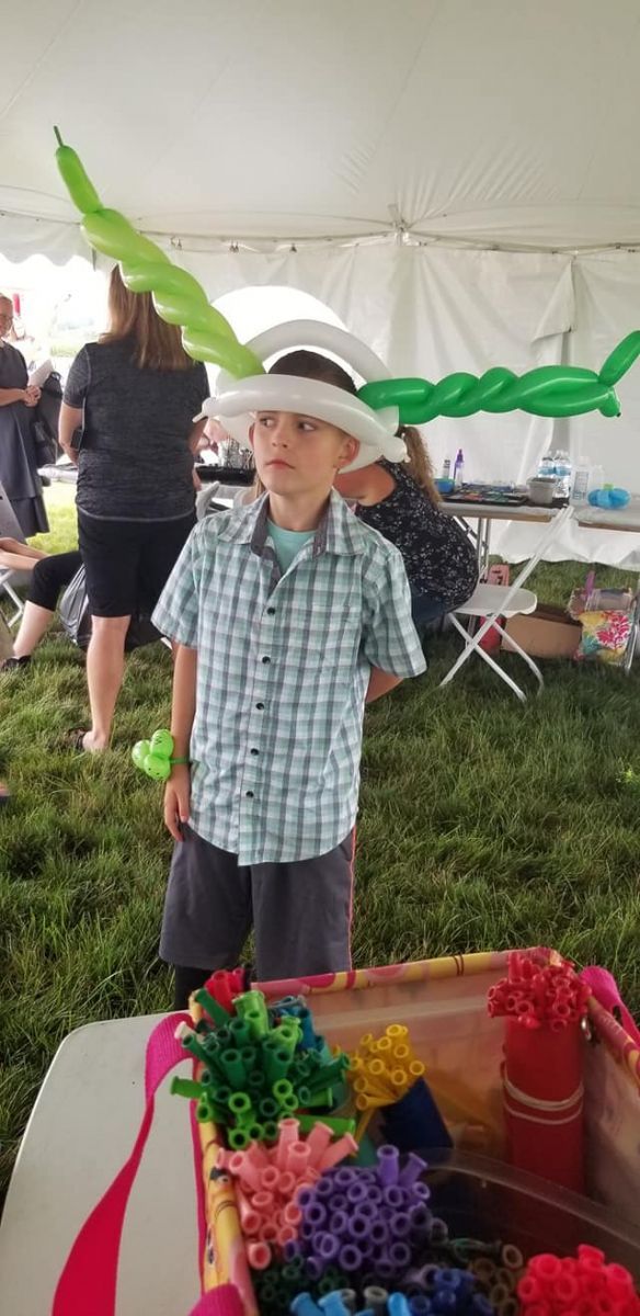 A young boy wearing a hat made of balloons is standing in front of a table.