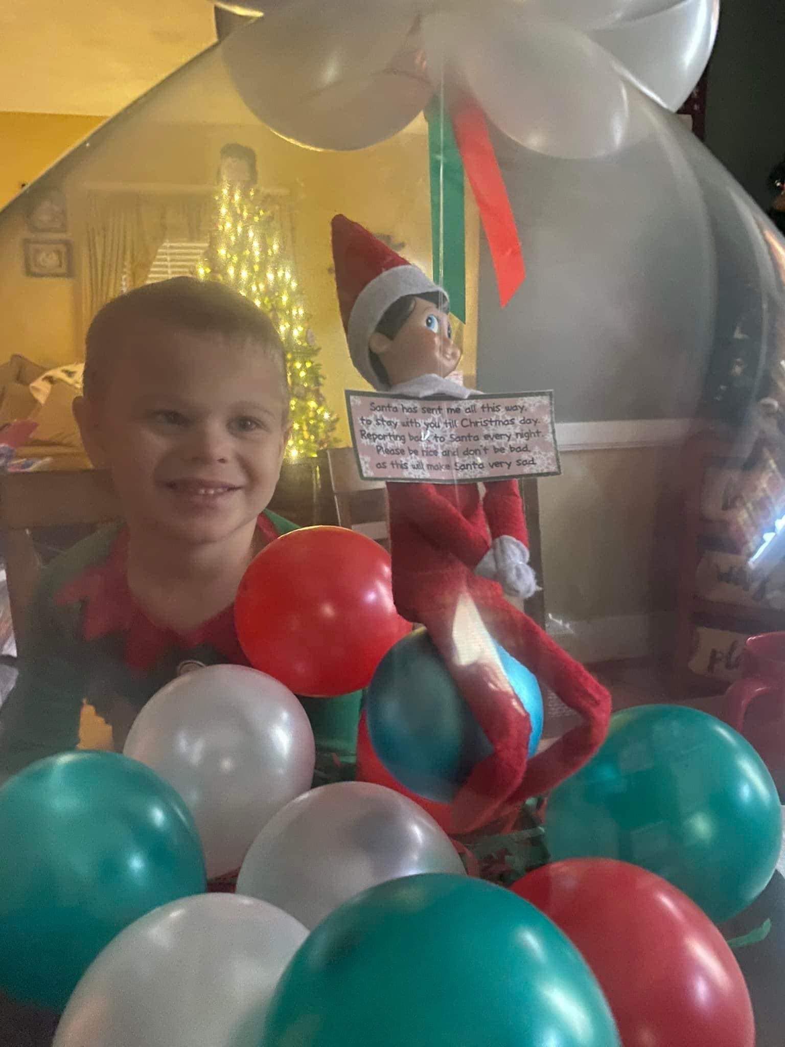 A little boy is standing in a pile of balloons next to an elf on the shelf.