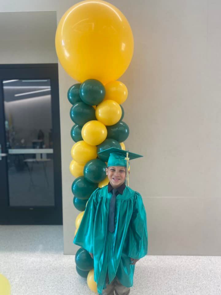 A young boy in a graduation cap and gown is standing in front of a balloon column