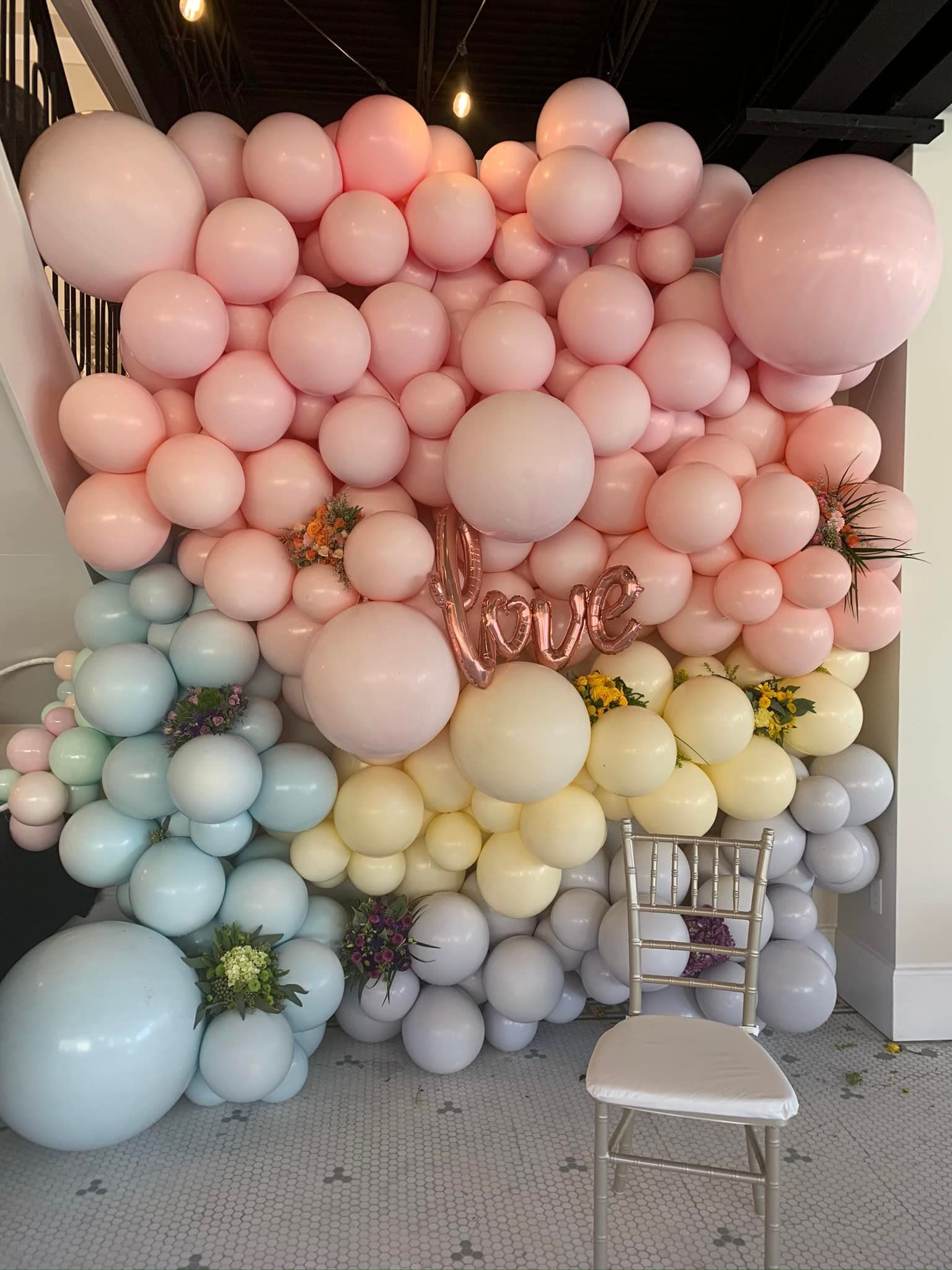 A wall of pink , yellow , and blue balloons with a chair in the foreground.