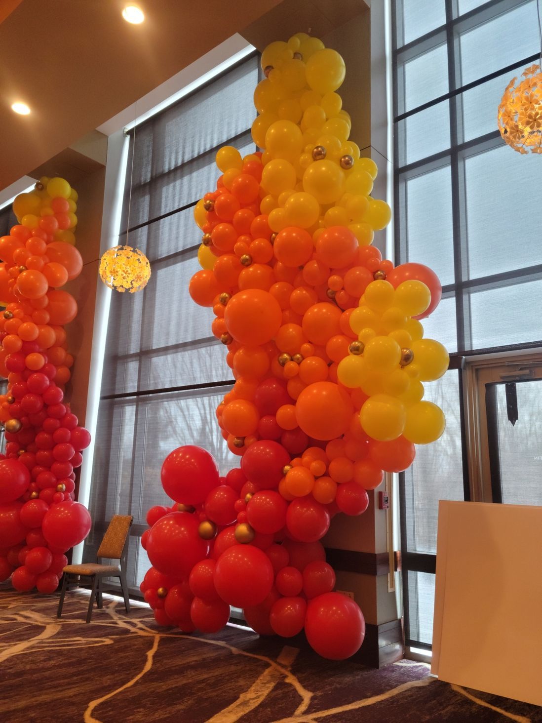 Colorful balloon arch in red, orange, and yellow hues, decorating a room near a window.