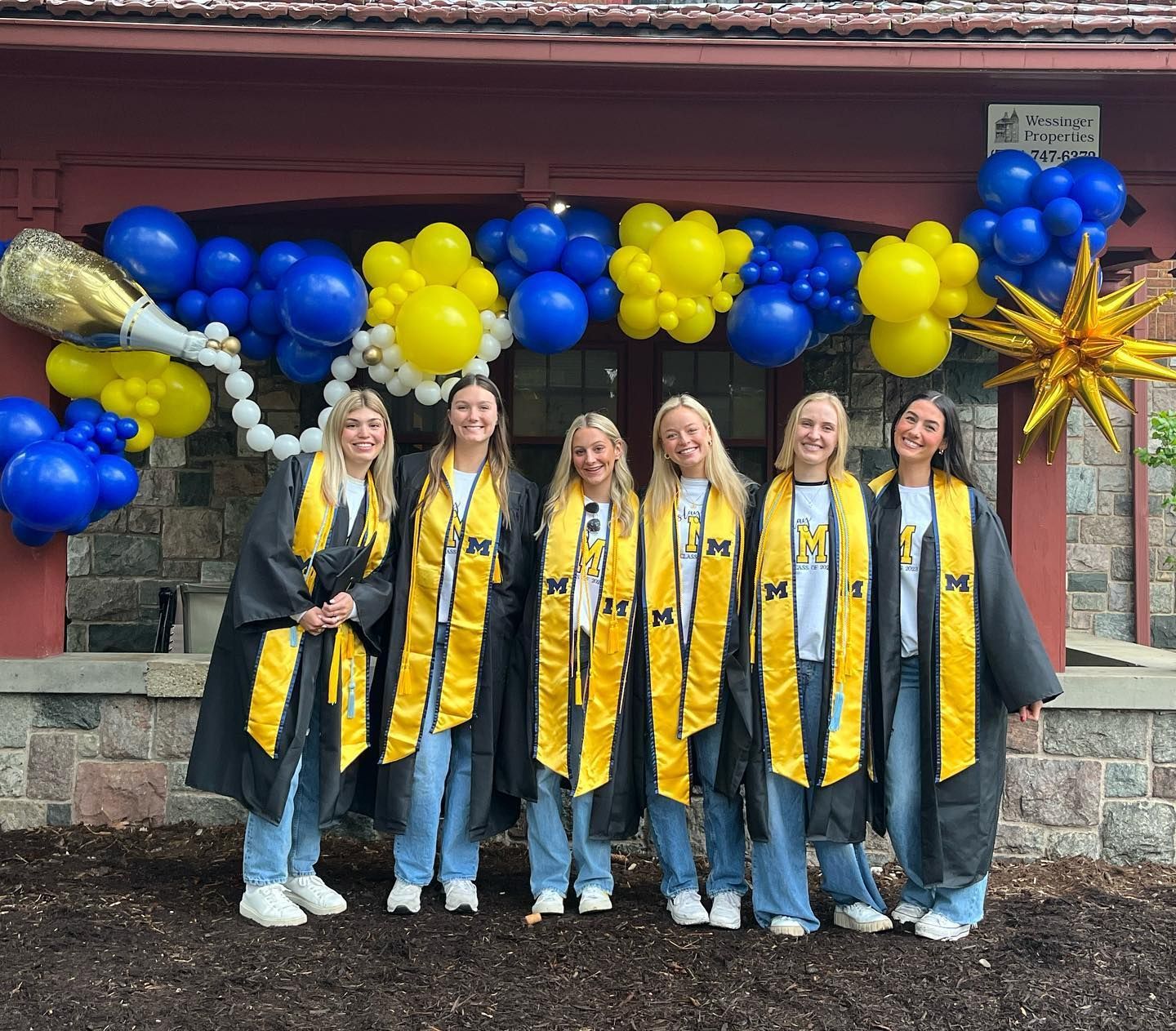A group of women in graduation gowns are standing in front of a building decorated with blue and yellow balloons.